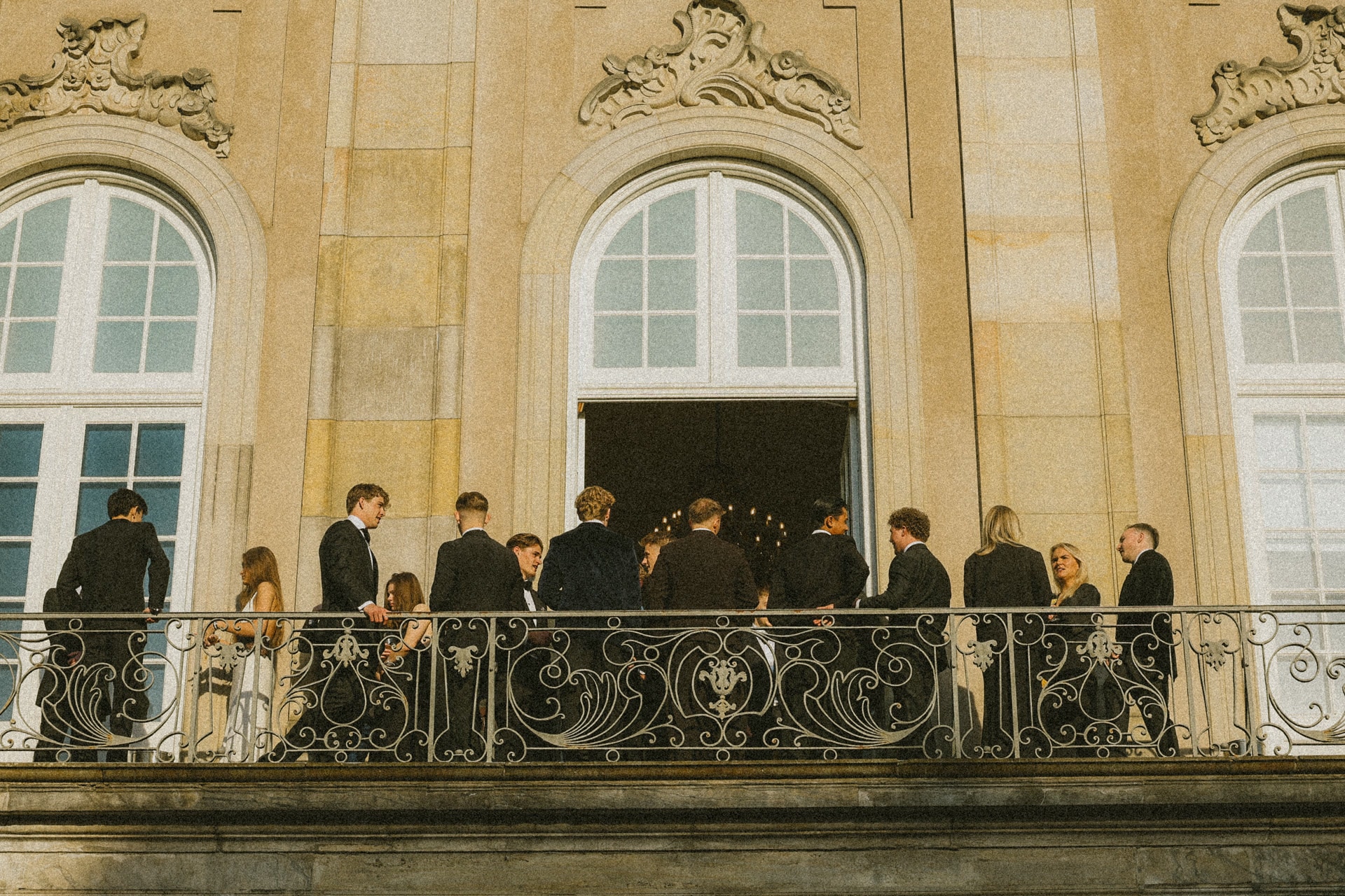 Wedding party in formal attire gathered on a wrought-iron balcony of a yellow townhouse at dusk