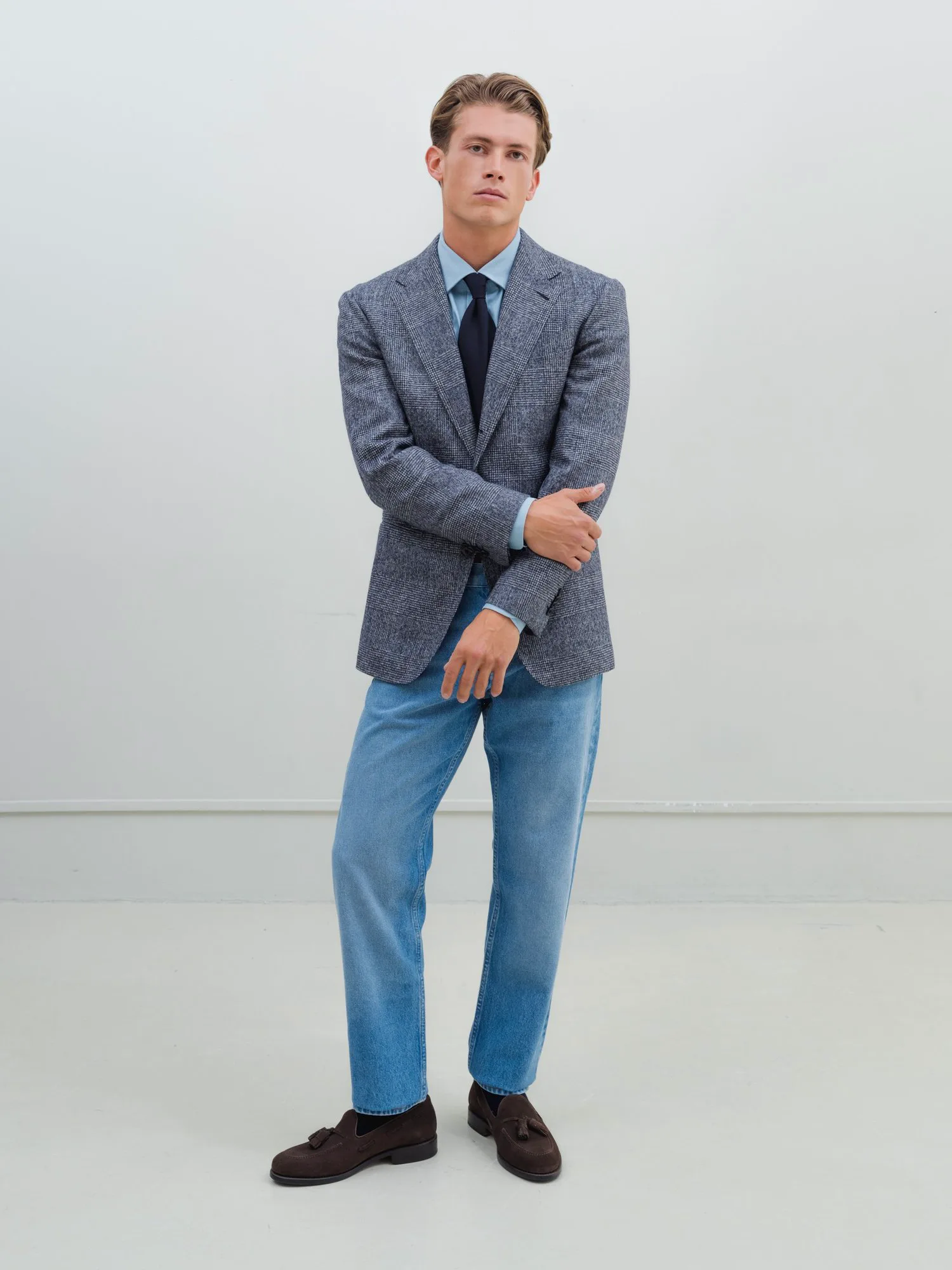 Man in a grey blazer, light blue shirt and light jeans in front of a light grey wall