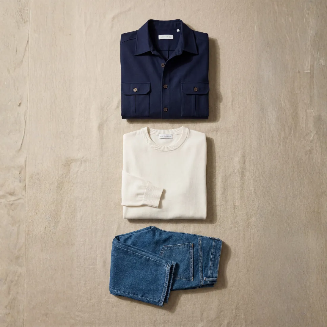 Navy overshirt, cream knit and blue jeans laid out flat side by side on a fabric surface
