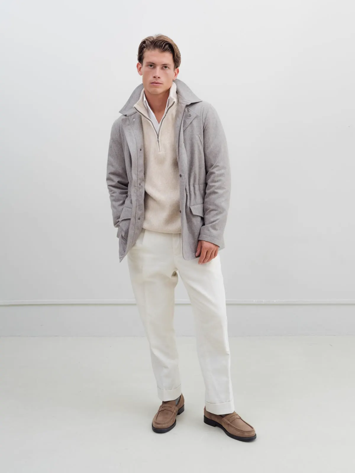 Man in a grey field jacket over a light knit and white trousers