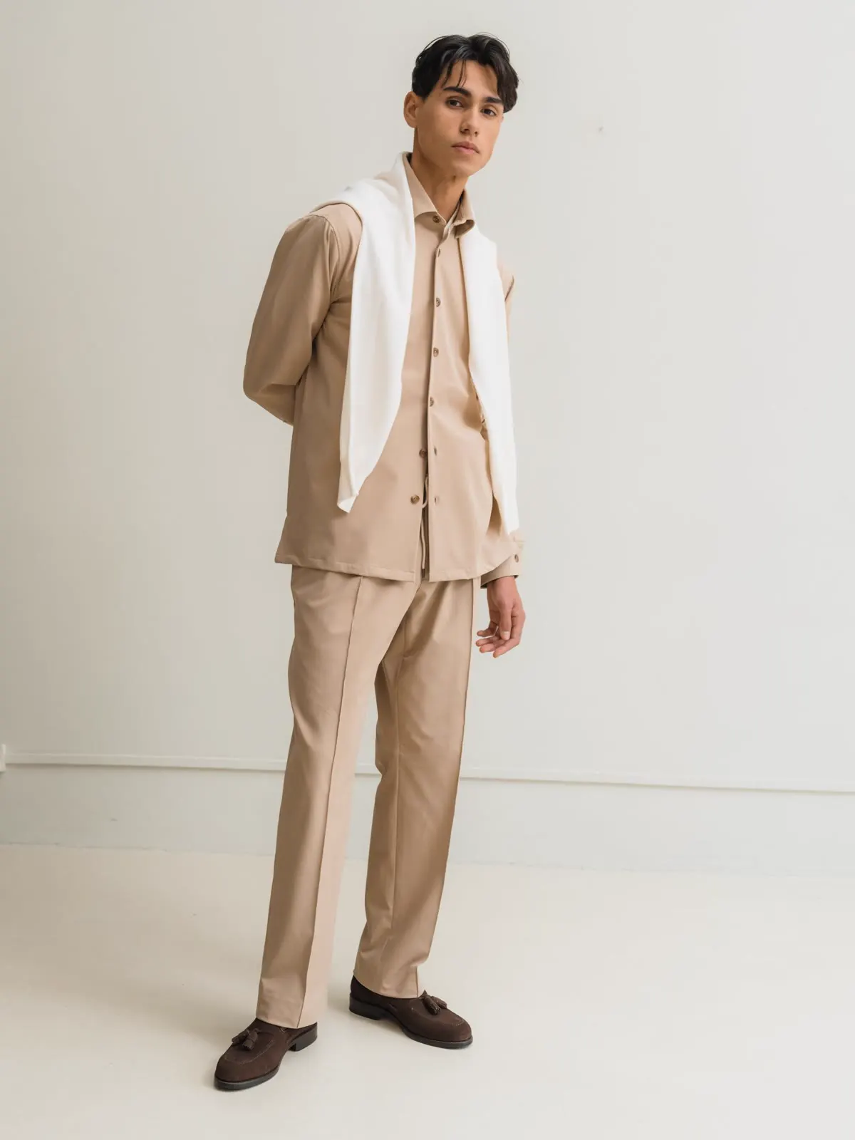 Man in a light brown overshirt and matching trousers with a white sweater over the shoulders