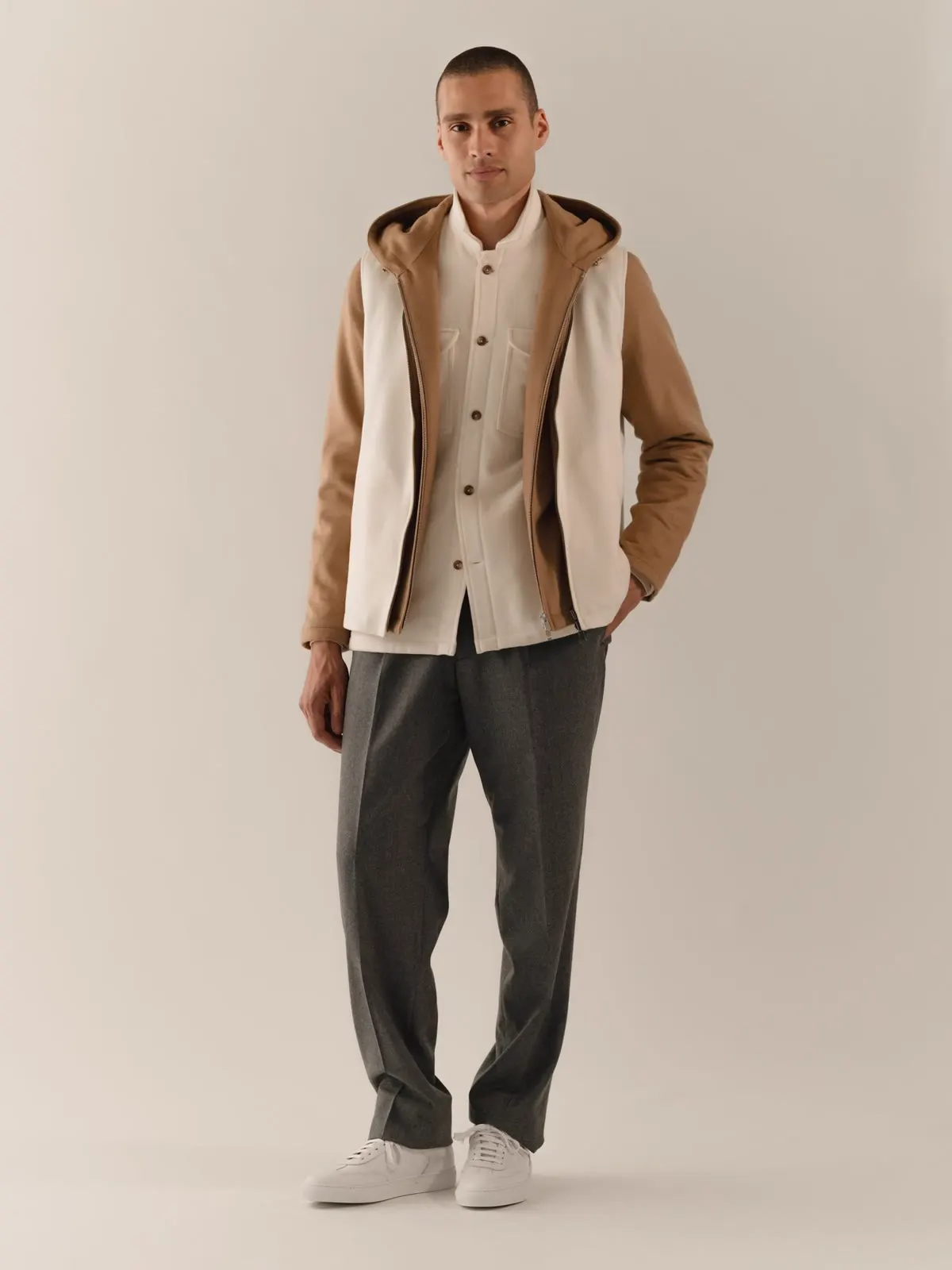 Man in a light overshirt under a brown hooded gilet, dark trousers and white shoes
