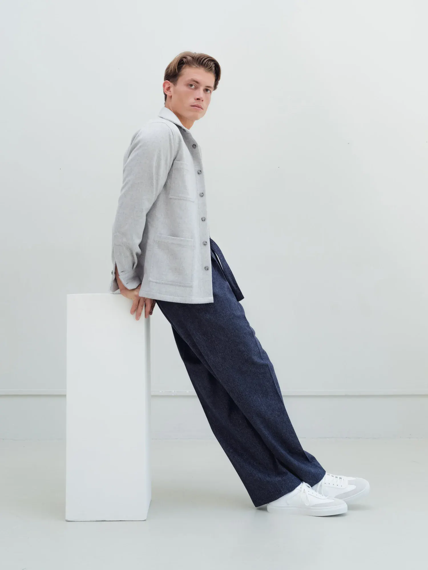 Man leaning against a white plinth in a light grey overshirt and dark blue trousers