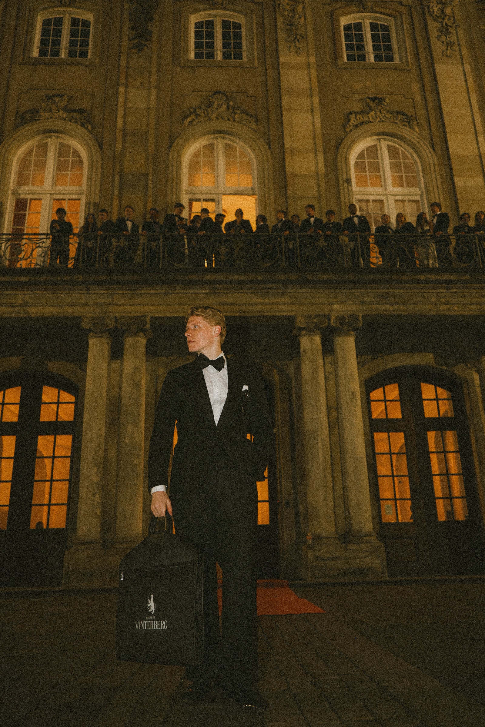 Man in a black tuxedo with bow tie holding a Vinterberg bag outside a lit townhouse at night